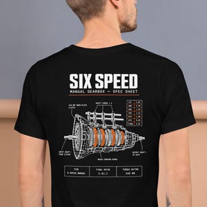 May include: A black t-shirt featuring a white and orange technical diagram of a six-speed manual gearbox. The graphic includes the text SIX SPEED MANUAL GEARBOX - SPEC SHEET, along with labels for components like shift forks and brass synchro rings.