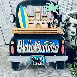 May include: Black wooden truck cutout with a beach theme. The truck bed has a sandcastle, palm tree, and surfboard. The back window says "Hello Summer" and the license plate says "Beach".