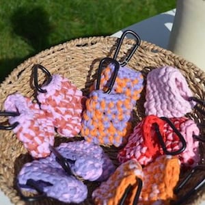 Könnte beinhalten: Eine Sammlung farbenfroher, gehäkelter Artikel mit schwarzen Karabinerhaken, präsentiert in einem Weidenkorb. Die Artikel weisen Farbtöne von Rosa, Orange, Lila und Rot auf. Der Korb steht auf einer weißen Oberfläche mit grünem Gras im Hintergrund.