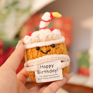 Puede incluir: Un cupcake de cumpleaños tejido a mano, con una cara sonriente, glaseado blanco y una vela roja y amarilla. El cupcake tiene una etiqueta que dice "Happy Birthday! You deserve all the best today and always!"