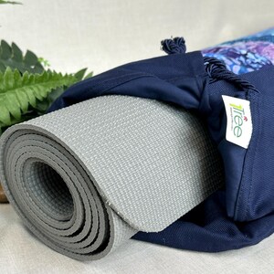 May include: A rolled-up gray yoga mat is partially visible inside a navy blue yoga mat bag. The bag has a drawstring closure and a decorative patterned section with blue and purple hues. A small white tag with a logo is attached.