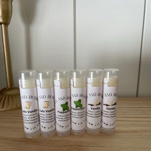 May include: Six clear tubes of lip balm with white labels. The labels have the words "AND ROSE" and the names of the flavors: "Cafe Vanilla", "Peppermint", and "Vanilla".