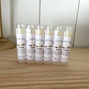 May include: Six white tubes of lip balm with a white label that says "Vanilla" and lists the ingredients. The label also says "Glow Lip" at the bottom.