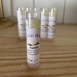 May include: A close-up of a vanilla flavored lip balm in a clear tube with a white label. The label reads "Vanilla" and lists the ingredients: Tallow, Castor Oil, Beeswax, Vitamin E, and Vanilla Oleoresin. The label also says "Slow Lip".