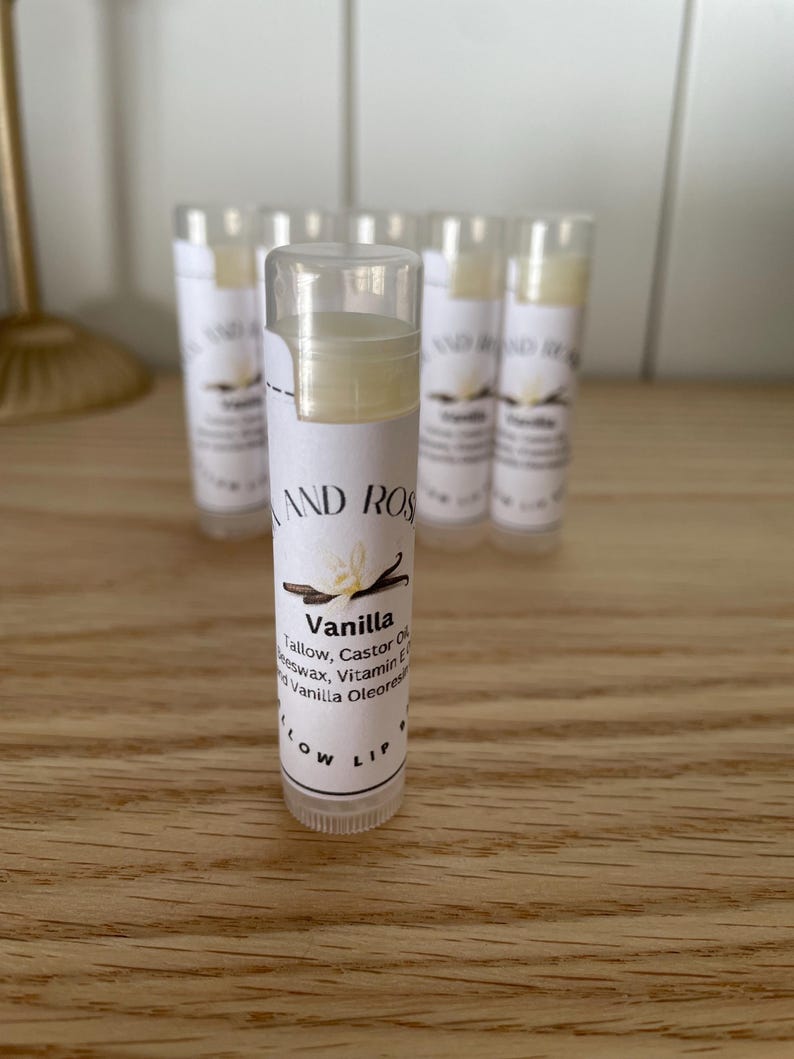 May include: A close-up of a vanilla flavored lip balm in a clear tube with a white label. The label reads "Vanilla" and lists the ingredients: Tallow, Castor Oil, Beeswax, Vitamin E, and Vanilla Oleoresin. The label also says "Slow Lip".