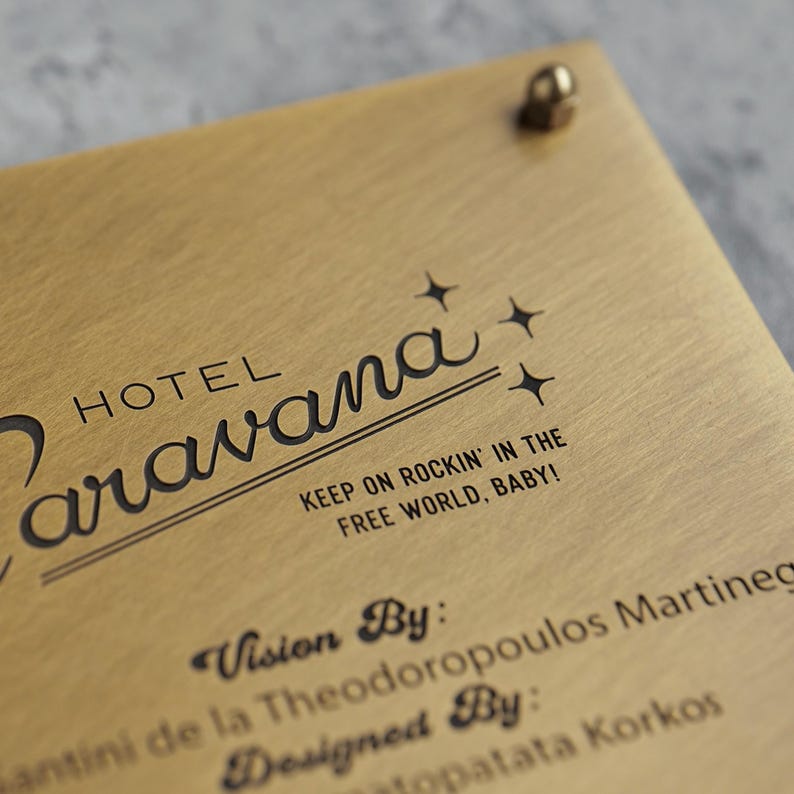 May include: A rectangular brushed gold metal plaque featuring the black text Hotel Caravana, the phrase Keep on Rockin' in the Free World, Baby!, and the names of the visionaries and designers. A small metallic fastener is visible at the top.