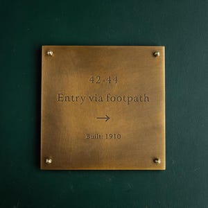 May include: A square brass plaque with a brushed finish, secured by four corner screws. The text reads 42-44, Entry via footpath, an arrow pointing right, and Built: 1910. The plaque is mounted on a dark green surface.