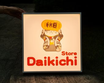 Op de afbeelding: Vierkant verlicht bord met een cartoonkat in traditionele Japanse kledij. Het bord toont het woord "Daikichi" in het rood, met "Store" erboven. De kat houdt een gouden munt met Japanse tekst vast.