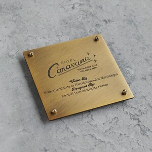 May include: A square brushed brass plaque with four small metal studs at the corners. The text reads Hotel Caravana, Keep on rockin in the free world, baby!, Vision By: Bobby Santini de la Theodoropoulos Martinegro, Designed By: Samuel Stamatopatata Korkos.