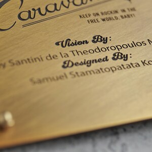 May include: A brushed gold metal plaque featuring black engraved text that reads Caravan, Keep on rockin in the free world, baby, Vision By: Santini de la Theodoropoulos M, and Designed By: Samuel Stamatopatata Ko, with a small gold rivet visible.