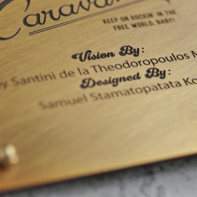 May include: A brushed gold metal plaque featuring black engraved text that reads Caravan, Keep on rockin in the free world, baby, Vision By: Santini de la Theodoropoulos M, and Designed By: Samuel Stamatopatata Ko, with a small gold rivet visible.