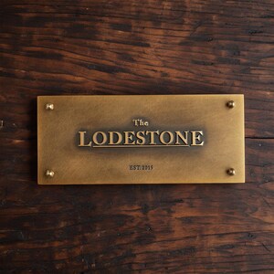 May include: A rectangular brass plaque with a brushed finish featuring the text "The LODESTONE" in raised lettering and "EST. 2015" below it. The plaque is secured at each of the four corners by small, rounded, metallic decorative fasteners.