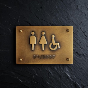 May include: A rectangular brass-toned metal sign featuring embossed icons of a person, a person in a dress, and a person in a wheelchair. The sign includes braille dots below the icons and is secured at each corner with a small metal screw.
