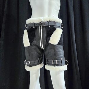 May include: Black leather shorts with white shearling accents and a black belt. The shorts feature a front zipper, buckle details, and a white stripe down the front. The shorts are displayed on a mannequin.