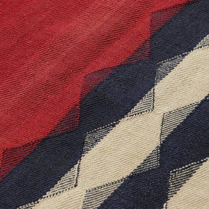 May include: Close-up of a woven textile with a geometric pattern. The design features a combination of red, navy blue, and beige diamond shapes. The texture of the fabric is visible, suggesting a soft, possibly natural material.