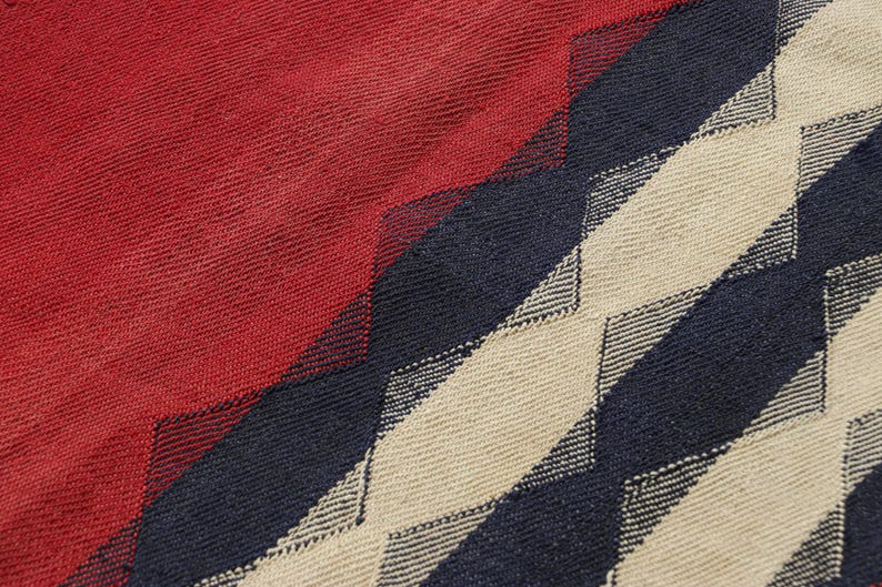 May include: Close-up of a woven textile with a geometric pattern. The design features a combination of red, navy blue, and beige diamond shapes. The texture of the fabric is visible, suggesting a soft, possibly natural material.