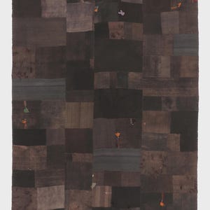 May include: A patchwork textile in shades of brown, black, and gray. The rectangular design is composed of various fabric squares, some with subtle variations in texture and tone. Small, colorful embellishments are scattered across the surface, adding a touch of detail.