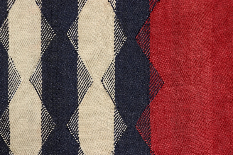 May include: Close-up of a woven fabric with a geometric pattern. The design features alternating vertical stripes of cream, navy blue, and red. The texture of the fabric is visible, suggesting a textured material.