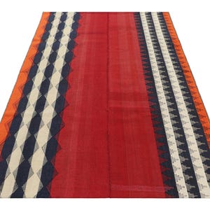 May include: A rectangular rug with a central red field and patterned borders. The borders feature geometric designs in navy, white, and orange. The rug is likely made of woven fabric and suitable for floor covering.