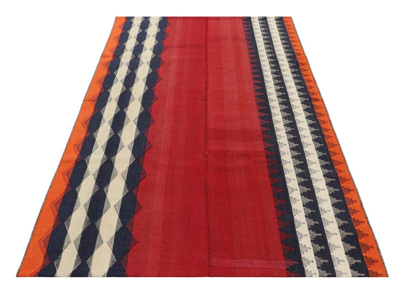 May include: A rectangular rug with a central red field and patterned borders. The borders feature geometric designs in navy, white, and orange. The rug is likely made of woven fabric and suitable for floor covering.