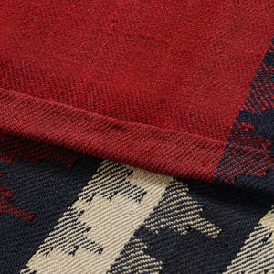 May include: Close-up of a red and plaid fabric. The fabric has a textured appearance with a deep red color dominating the upper portion. The lower right corner features a pattern of navy blue and beige geometric shapes.