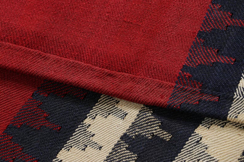 May include: Close-up of a red and plaid fabric. The fabric has a textured appearance with a deep red color dominating the upper portion. The lower right corner features a pattern of navy blue and beige geometric shapes.