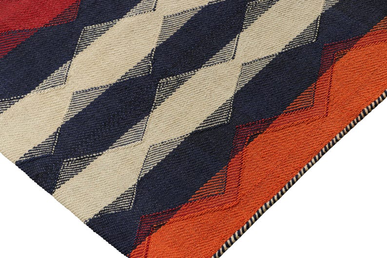 May include: Close-up of a woven rug with a geometric pattern. The rug features alternating diagonal stripes of navy blue, cream, and orange. The edges are finished with a black and white border. The rug is made of a textured material.