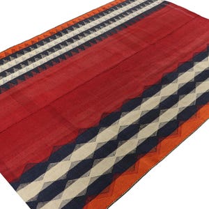 May include: A rectangular red rug with geometric patterns. The rug features bands of orange, white, navy blue, and black triangles and stripes. The rug is made of woven fabric and has a textured appearance.