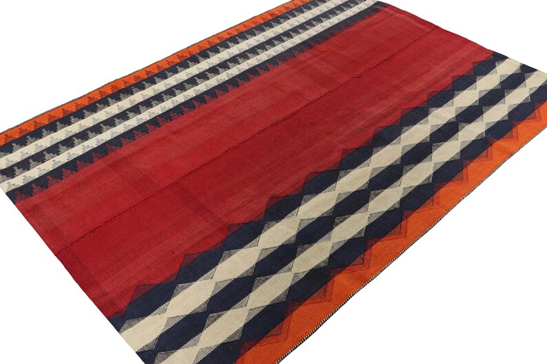May include: A rectangular red rug with geometric patterns. The rug features bands of orange, white, navy blue, and black triangles and stripes. The rug is made of woven fabric and has a textured appearance.
