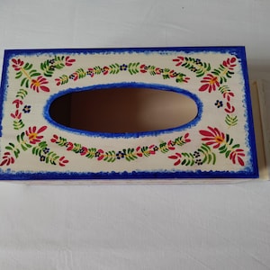 May include: Wooden tissue box, rectangular with a cream base, blue border, and oval opening. It features a floral design in red, green, and blue, offering a rustic aesthetic. The box is approximately 25 cm long.