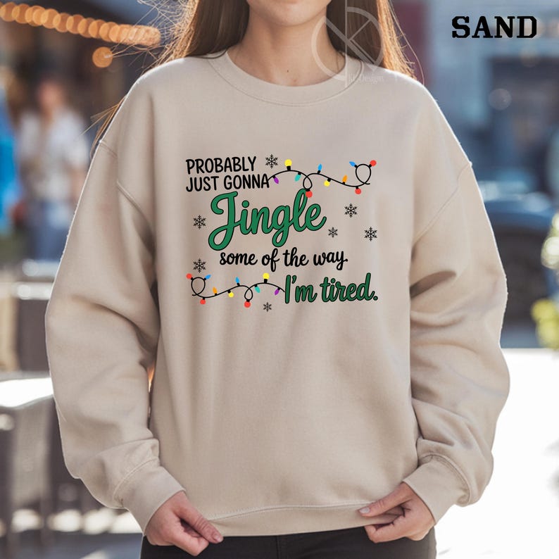 May include: A sand-colored sweatshirt with the text "PROBABLY JUST GONNA Jingle some of the way I'm tired." The text is in green and black, with snowflake and string light graphics. The word "SAND" is in the upper right corner.