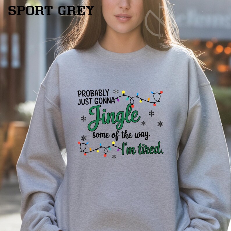 May include: A light gray sweatshirt with the text "Probably Just Gonna Jingle some of the way I'm tired." The text is in green and black with snowflake and Christmas light accents. The sweatshirt is labeled "SPORT GREY".