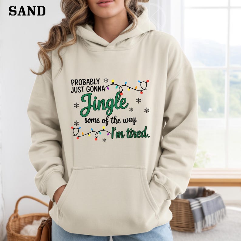 May include: Sand-colored hoodie with a front pocket and a drawstring hood. The front features the text "PROBABLY JUST GONNA Jingle some of the way. I'm tired." in green and black, with snowflake and Christmas light accents.