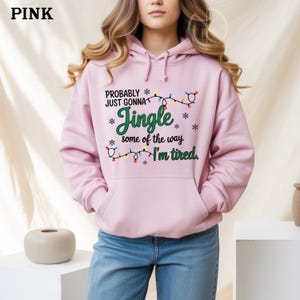 May include: A pink hooded sweatshirt with a front pocket and the text "PROBABLY JUST GONNA Jingle some of the way I'm tired." The text is accompanied by snowflake and Christmas light graphics. The word "PINK" is in the upper left corner.
