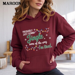 May include: Maroon hoodie with a front pocket and a drawstring hood. The hoodie features the text "Probably Just Gonna Jingle some of the way. I'm tired." in white and green, with snowflake and Christmas light graphics.