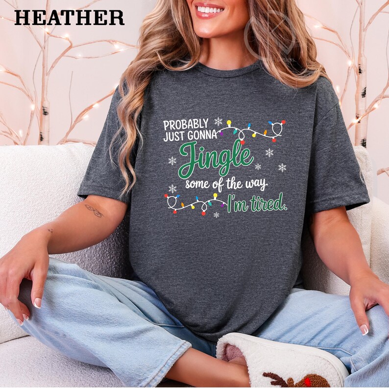 May include: Dark gray heather t-shirt with the text "PROBABLY JUST GONNA Jingle some of the way. I'm tired." The text is in green and white with a string of Christmas lights. The word "HEATHER" is at the top.