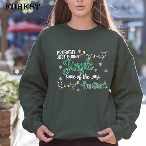 May include: Forest green sweatshirt with the text "PROBABLY JUST GONNA Jingle some of the way I'm tired." The text is in white and green with snowflake and Christmas light accents. The sweatshirt is a crew neck style.
