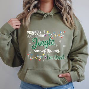 May include: An olive green hooded sweatshirt with the text "Probably just gonna Jingle some of the way, I'm tired." The design includes snowflakes and a string of Christmas lights in various colors. The sweatshirt has a front pocket.