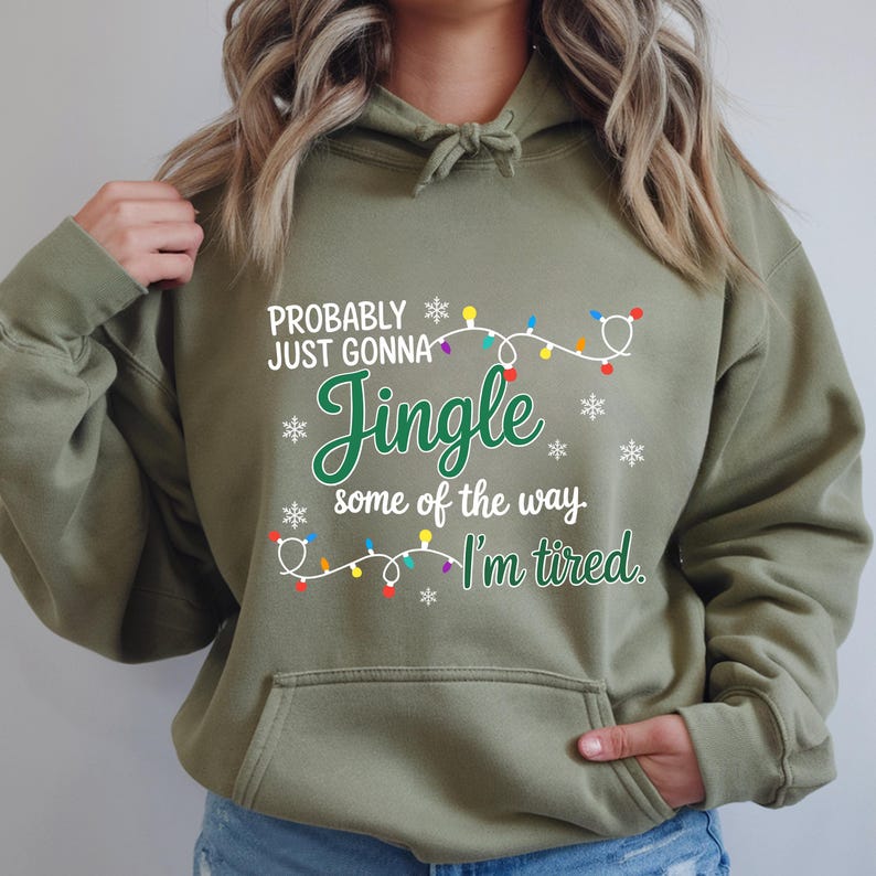 May include: An olive green hooded sweatshirt with the text "Probably just gonna Jingle some of the way, I'm tired." The design includes snowflakes and a string of Christmas lights in various colors. The sweatshirt has a front pocket.