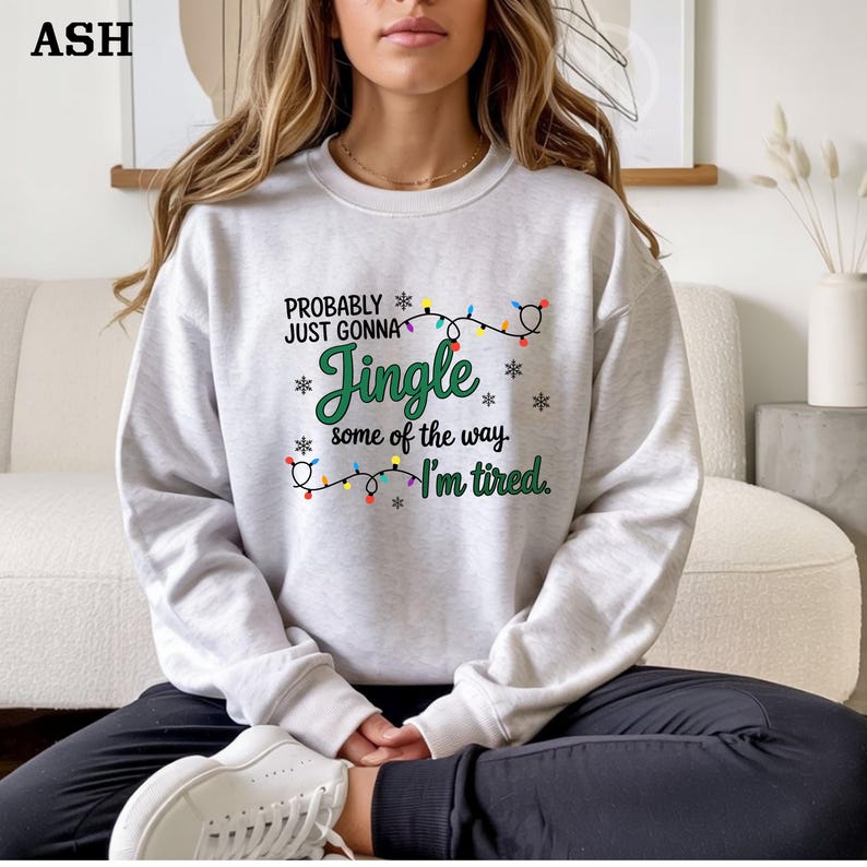 May include: A light gray sweatshirt with the text "PROBABLY JUST GONNA Jingle some of the way I'm tired." The text is in black and green with snowflake and Christmas light accents. The sweatshirt is a crew neck style.