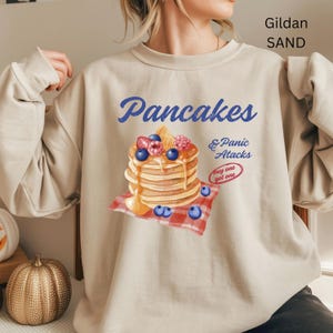 May include: Sand-colored sweatshirt with the words "Pancakes & Panic Attacks" in blue script. A stack of pancakes with syrup, blueberries, and raspberries is printed on the front. The sweatshirt is a Gildan brand.
