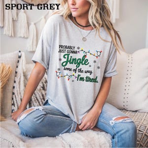 May include: Sport grey t-shirt with the text "Probably Just Gonna Jingle some of the way, I'm tired." The text is in green and black with a string of Christmas lights. The shirt is a short-sleeved, casual style.