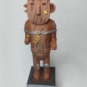 Könnte beinhalten: Braune Figur einer Stammesfigur mit rechteckigem Kopf und Holzstruktur. Die Figur hat goldene und silberne Akzente, darunter Bandagen und Armbänder. Der Sockel hat eingravierten Text.