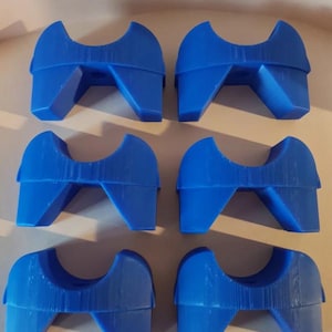 Può includere: Sei oggetti blu di forma geometrica sono disposti in due file da tre. Ogni oggetto ha una parte superiore curva e due gambe angolate, che assomigliano a una sella o un supporto stilizzato. Gli oggetti sono di un blu vibrante e uniforme.