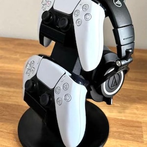 Peut inclure: Un support noir avec deux manettes blanches PlayStation 5 et un casque noir avec le logo Audio-Technica sur les écouteurs.