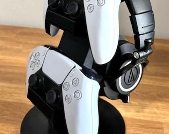 May include: A black stand with two white PlayStation 5 controllers and a black pair of headphones with the Audio-Technica logo on the earcups.