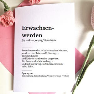 May include: A white card with the German word "Erwachsenwerden" printed in black, meaning "to mature". The card is inside a pink envelope, with crocheted pink flowers in the background.