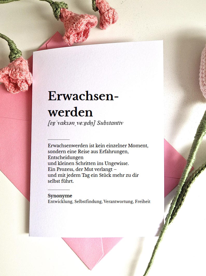 May include: A white card with the German word "Erwachsenwerden" printed in black, meaning "to mature". The card is inside a pink envelope, with crocheted pink flowers in the background.