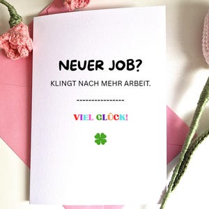 May include: A white greeting card with the text "NEUER JOB? KLINGT NACH MEHR ARBEIT." in black. Below, the words "VIEL GLÜCK!" are in rainbow colours, and a green four-leaf clover is at the bottom. The card is inside a pink envelope.