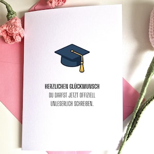Könnte beinhalten: Eine weiße Glückwunschkarte zur Graduierung mit einer Illustration eines marineblauen Mortarboards und einer goldenen Quaste. Die Karte befindet sich in einem rosa Umschlag. Rosa gehäkelte Blumen im Hintergrund. Deutscher Text.